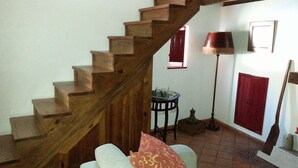 Un escalier en bois menant à l’étage supérieur, une table ronde avec des pièces d’échecs, une porte rouge et un lampadaire.