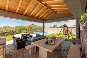 An outdoor patio with stone flooring, a wooden ceiling, a dining table with a bottle and glasses, and a view of a swimming pool and a children's play structure. - Villa Elder ZadarVillas (Lepuri)