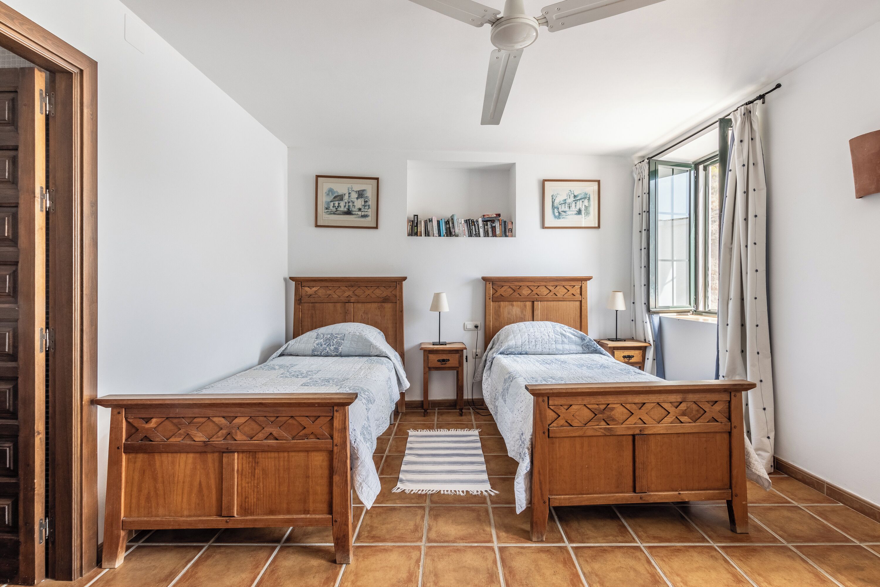 Une chambre avec deux lits en bois, un ventilateur de plafond, une fenêtre avec des rideaux et une bibliothèque.
