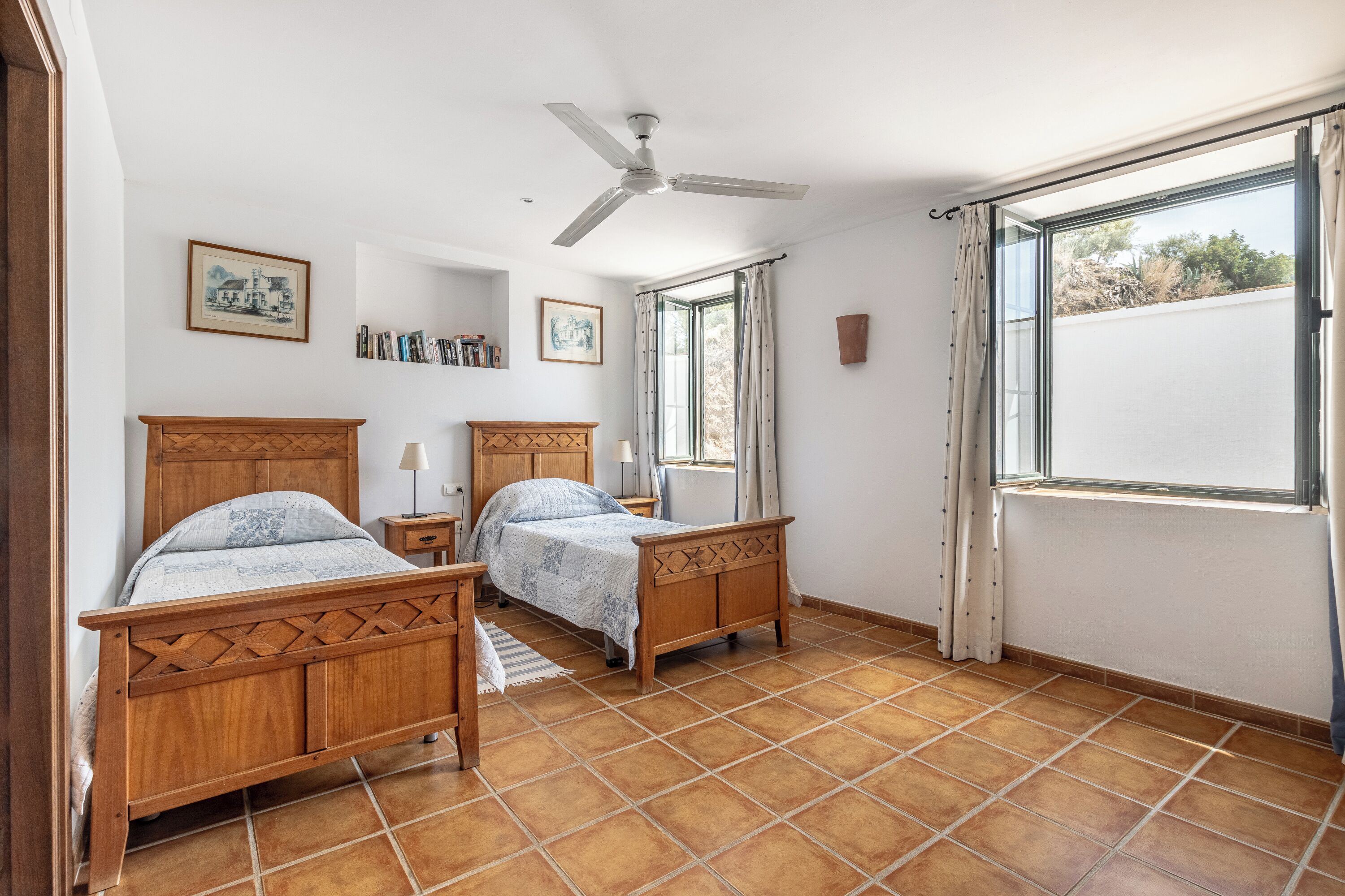Une chambre avec deux lits en bois, un ventilateur de plafond, une fenêtre avec des rideaux et un sol carrelé.