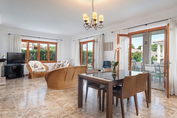 A spacious living area with a dining table, chairs, a TV, wicker furniture, and large windows offering a view of the outdoors.