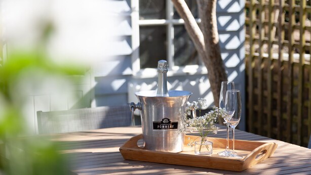 Une table en bois, un seau à champagne en métal, deux verres et un petit vase avec des fleurs.