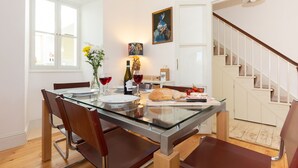Un coin repas avec une table en verre, des chaises, un vase de fleurs, une bouteille de vin et une miche de pain.