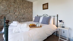 Un lit bien fait, avec du linge de lit blanc, un plateau de petit-déjeuner comprenant des croissants et des fruits, une table de chevet avec une lampe et un mur en pierre.