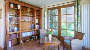Una habitación con una amplia estantería de madera, una silla de mimbre, una mesa con un florero y un gran ventanal con cortinas.