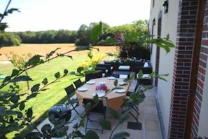 Un espace extérieur aménagé pour les repas, avec une table dressée pour quatre personnes, entourée de chaises, et offrant une vue sur un champ verdoyant et des arbres en arrière-plan.