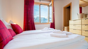 A hotel room with a bed, red curtains, a wooden dresser, and a view of snow-covered mountains through the window.