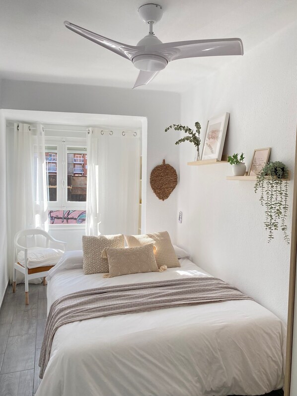 A bedroom with a bed, a ceiling fan, a window with curtains, a chair, and wall decorations including a hanging plant and framed pictures.