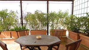 Um pátio externo com uma mesa redonda e seis cadeiras de madeira, rodeado por plantas em vasos.
