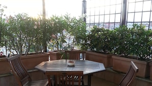 Uma varanda coberta com uma mesa redonda e quatro cadeiras de madeira, cercada por plantas em vasos.