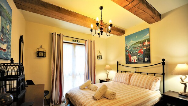 A neatly made bed with striped bedding and rolled towels, a chandelier, a painting of a coastal scene, and a wooden ceiling beam. - Lobster Pot in Padstow Is A 'fisherman's Style' Cottage With A Parking Space. (Padstow)