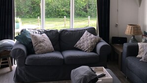A living room with a dark grey sofa, a matching ottoman, and a small side table with a lamp. Large windows offer a view of a green garden and a white building.