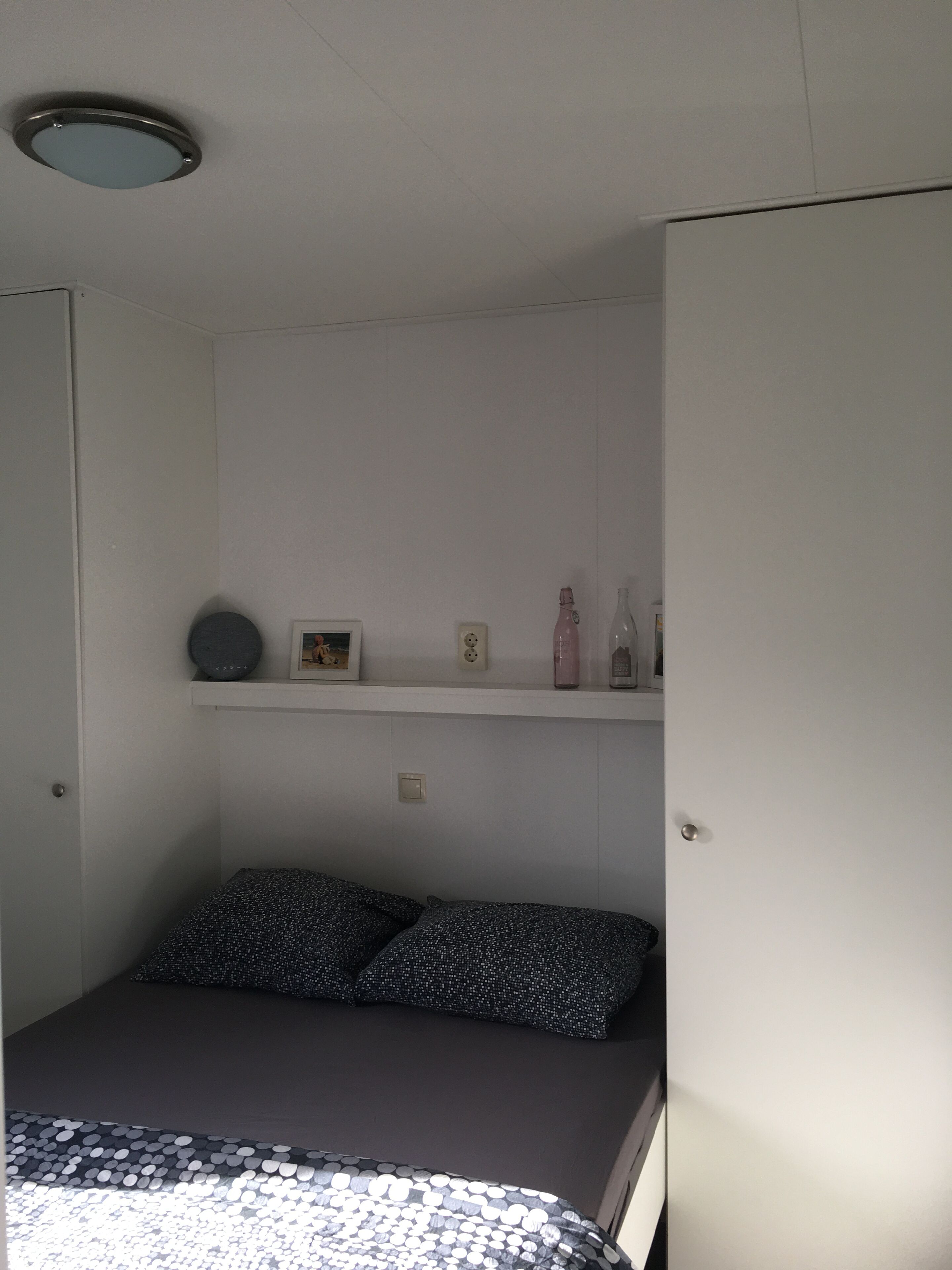 Ein kleines Zimmer mit einem Einzelbett, einer weißen Wand, einem Regal mit persönlichen Gegenständen und einer Deckenleuchte.