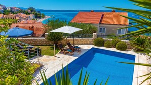 Une piscine entourée de chaises longues et d’un parasol, bordée de murs de pierre et de verdure, avec vue sur la mer et les bâtiments en arrière-plan.