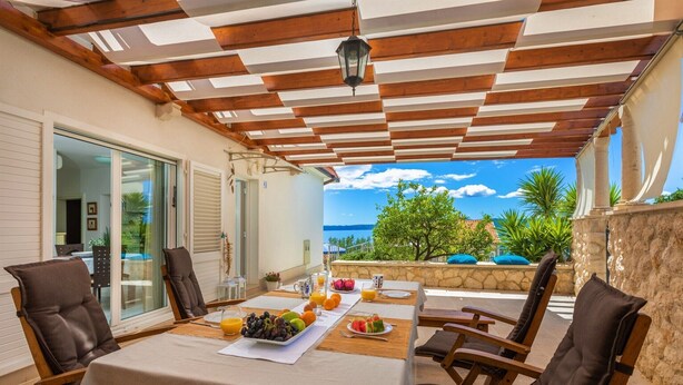 Un espace de restauration en plein air avec une table dressée pour le petit-déjeuner, comprenant des fruits et des boissons, sous une pergola en bois offrant une vue sur la mer et la verdure.