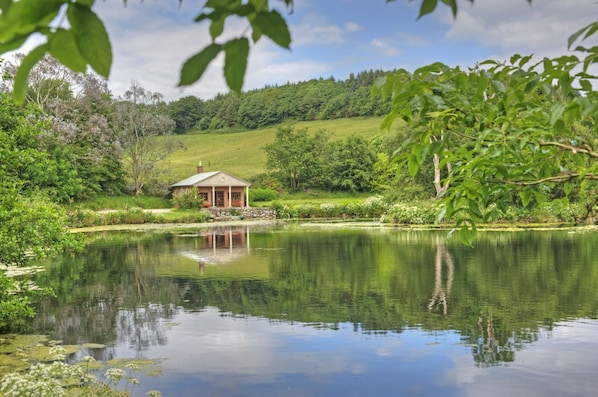 A tranquil pond with a small gazebo, surrounded by lush greenery and a hill in the background.