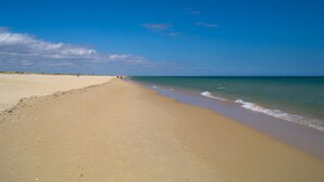 A wide sandy beach with gentle waves, a clear blue sky, and a few people walking along the shore. - Lovingly renovated Fisherman's cottage « Luazinha » (Tavira)