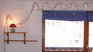 Ein gemütliches Zimmer mit einem hölzernen Bett, einer bunten Steudecke, einem kleinen Tisch mit Lampe und einem Fenster mit Vorhängen.
