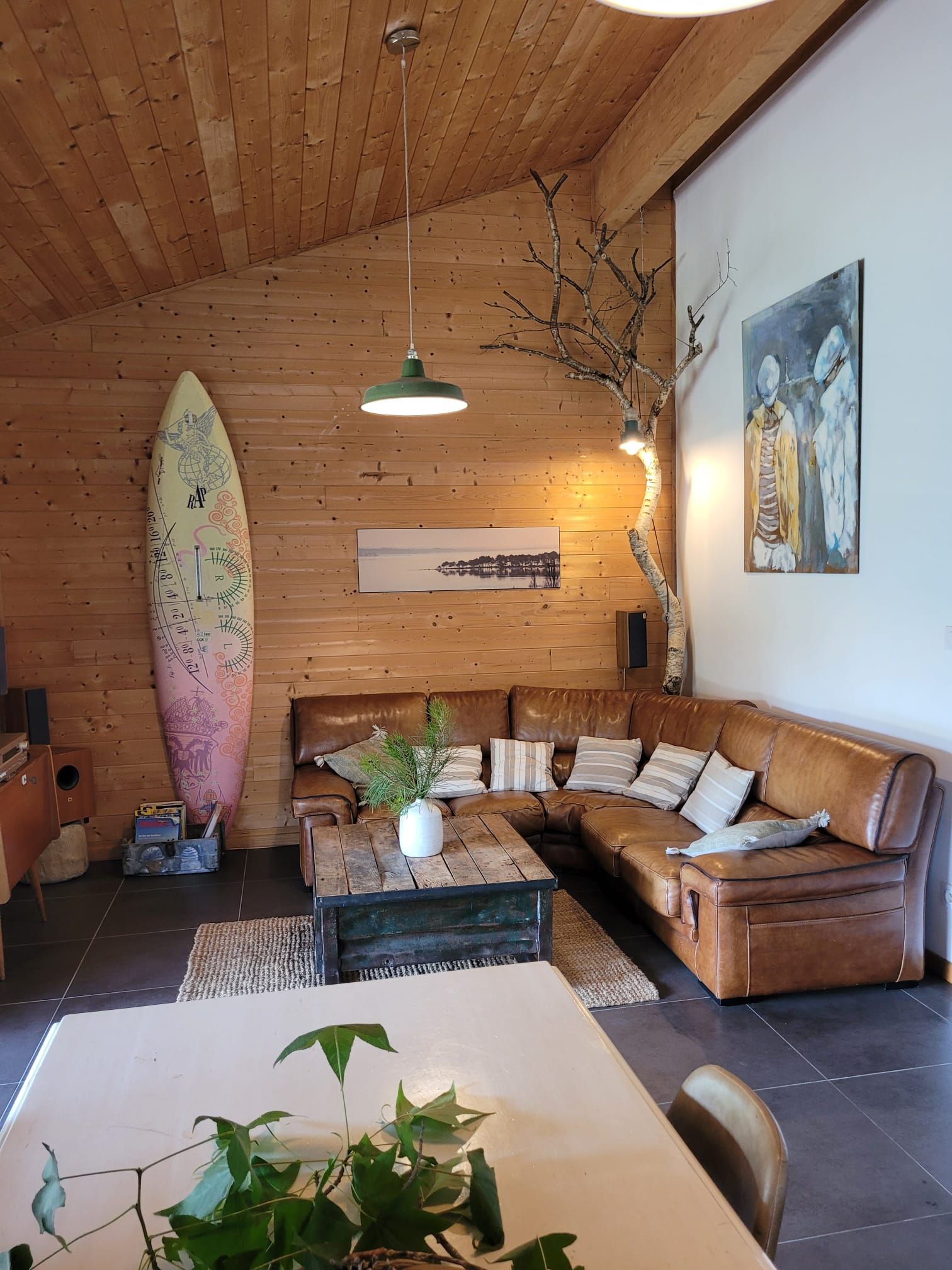 A cozy living room with a wooden ceiling, a large surfboard, a sofa, a coffee table, and a painting on the wall.