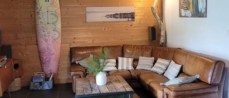 A cozy living room with a wooden ceiling, a large surfboard, a sofa, a coffee table, and a painting on the wall.