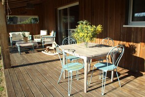 An outdoor wooden deck with a table and chairs, a potted plant, and a sliding glass door. - Wooden house by the sea (Hourtin)