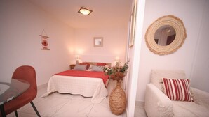 A bedroom with a bed, bedside tables, a chair, a small table, and a mirror on the wall. - Nice studio, spacious, air-conditioned, near village, sea view in small residence. (Cargèse)