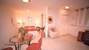 A cozy room with a bed, a dining table with chairs, a white armchair, and a small table with a vase. - Nice studio, spacious, air-conditioned, near village, sea view in small residence. (Cargèse)