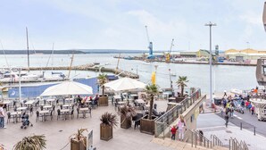 Ein Café am Wasser mit Außenplätzen, Blick auf den Yachthafen und ein weiter Industriebezirk im Hintergrund.