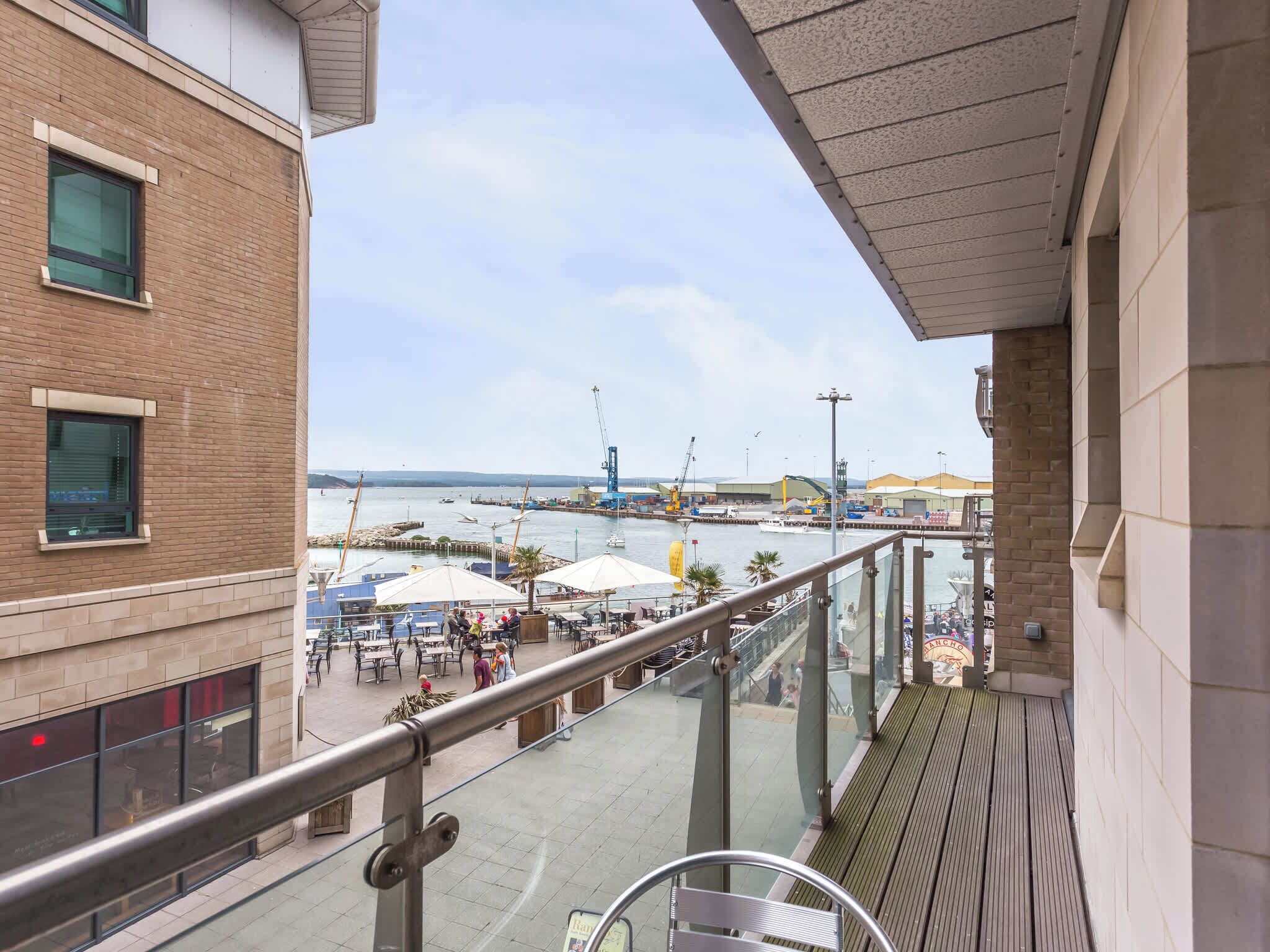 Un balcon donnant sur un port, des sièges d’extérieur et un bâtiment doté de grandes fenêtres.