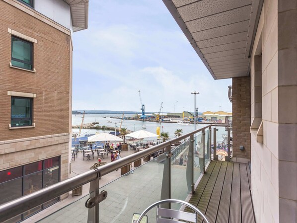 Ein Balkon mit Blick auf einen Hafen, Sitzgelegenheiten im Freien und ein Gebäude mit großen Fenstern.