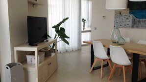 A modern living room with a TV, a dining table with chairs, a large plant, and a window with sheer curtains. - BALMINS APARTMENT, FULLY EQUIPPED 50 MTS FROM THE BEACH OF ST.SEBASTIA (SITGES)