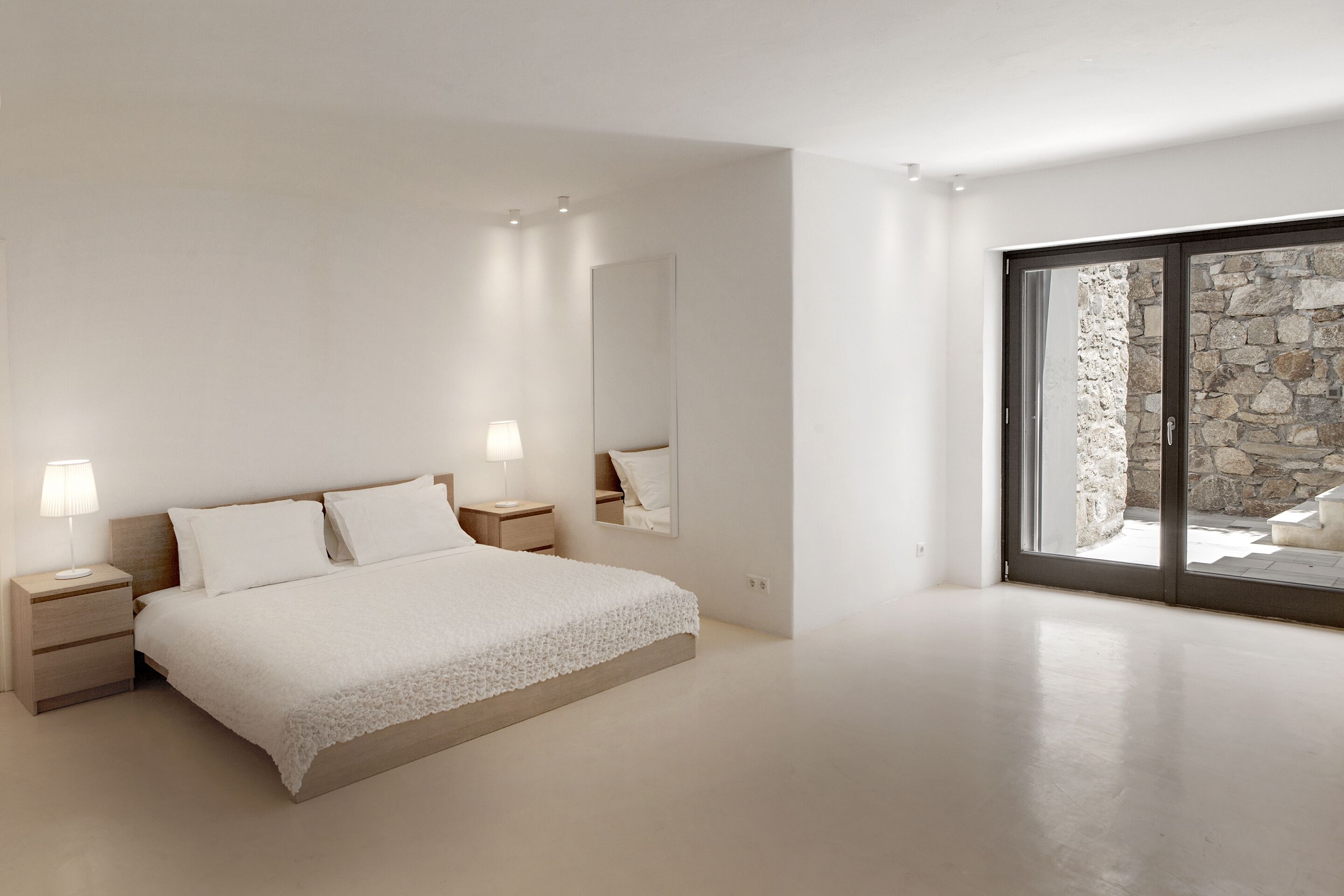 Une chambre moderne avec un grand lit, des tables de chevet avec des lampes, un miroir et une vue sur un mur en pierre, visible à travers une porte en verre.