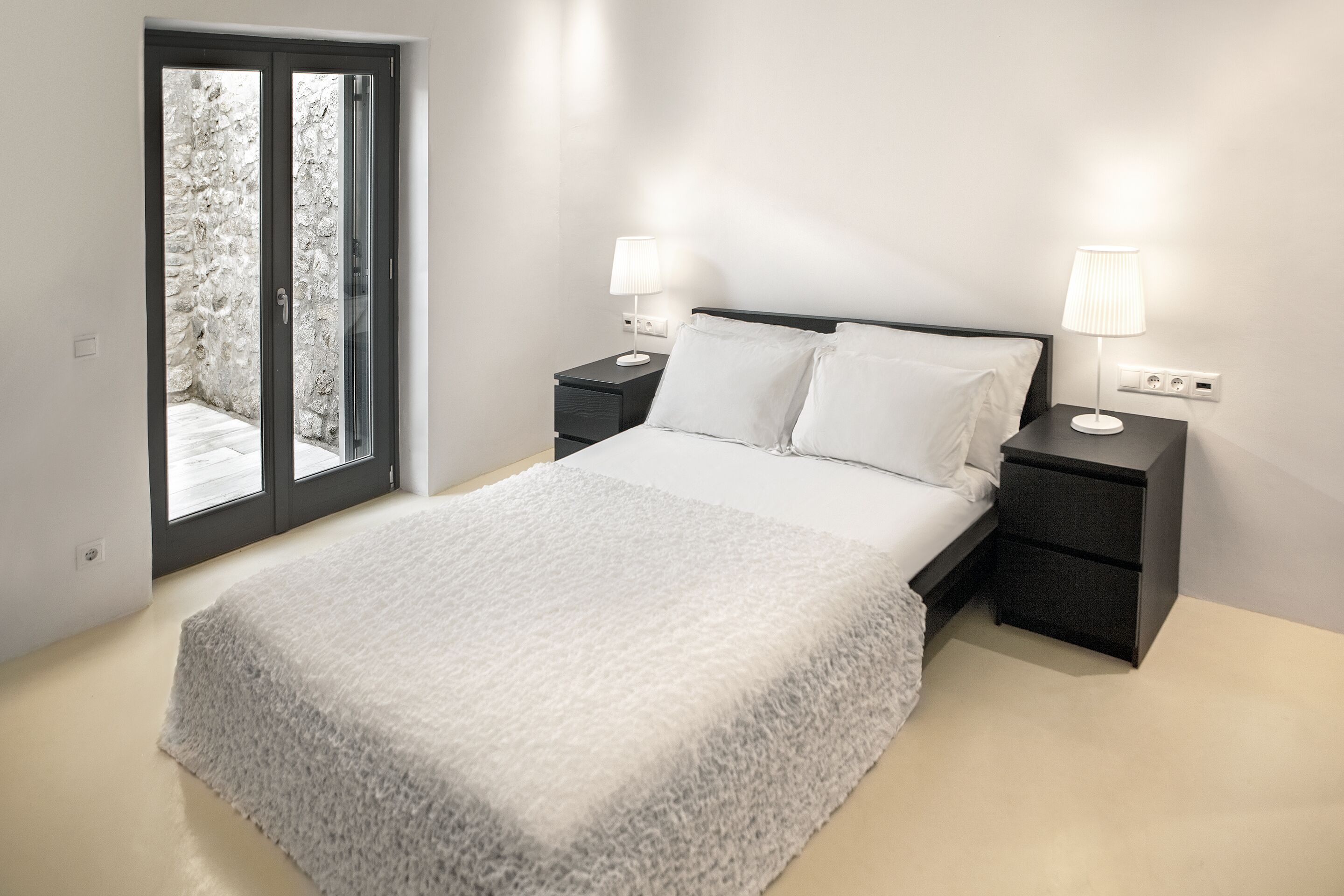 Une chambre à coucher avec un lit, deux tables de chevet avec des lampes, une grande fenêtre donnant sur un mur en pierre et une porte.
