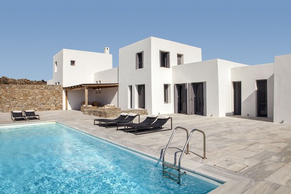 A modern pool area with a clear blue pool, black lounge chairs, and a stone wall. - New Liberation Villas (Mykonos)