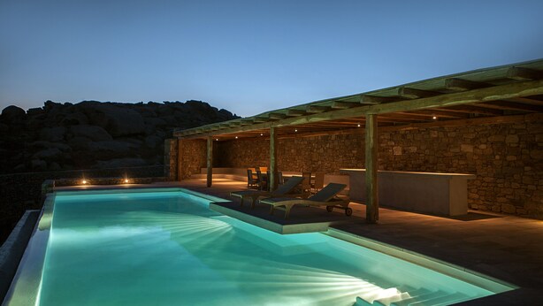 A swimming pool with a stone wall and lounge chairs. - New Liberation Villas (Mykonos)