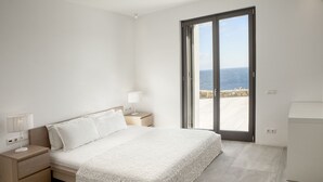 A bedroom with a bed, nightstands, a lamp, and a view of the sea through a sliding door. - New Liberation Villas (Mykonos)