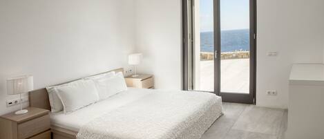 A bedroom with a bed, nightstands, a lamp, and a view of the sea through a sliding door.