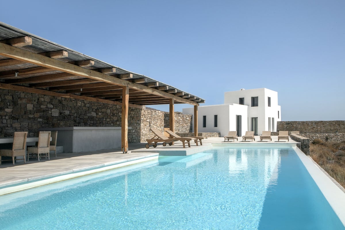 A modern pool area with stone walls, wooden pergola, and lounge chairs.