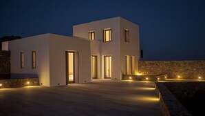 A modern two-story building with illuminated windows and a stone wall. - New Liberation Villas (Mykonos)