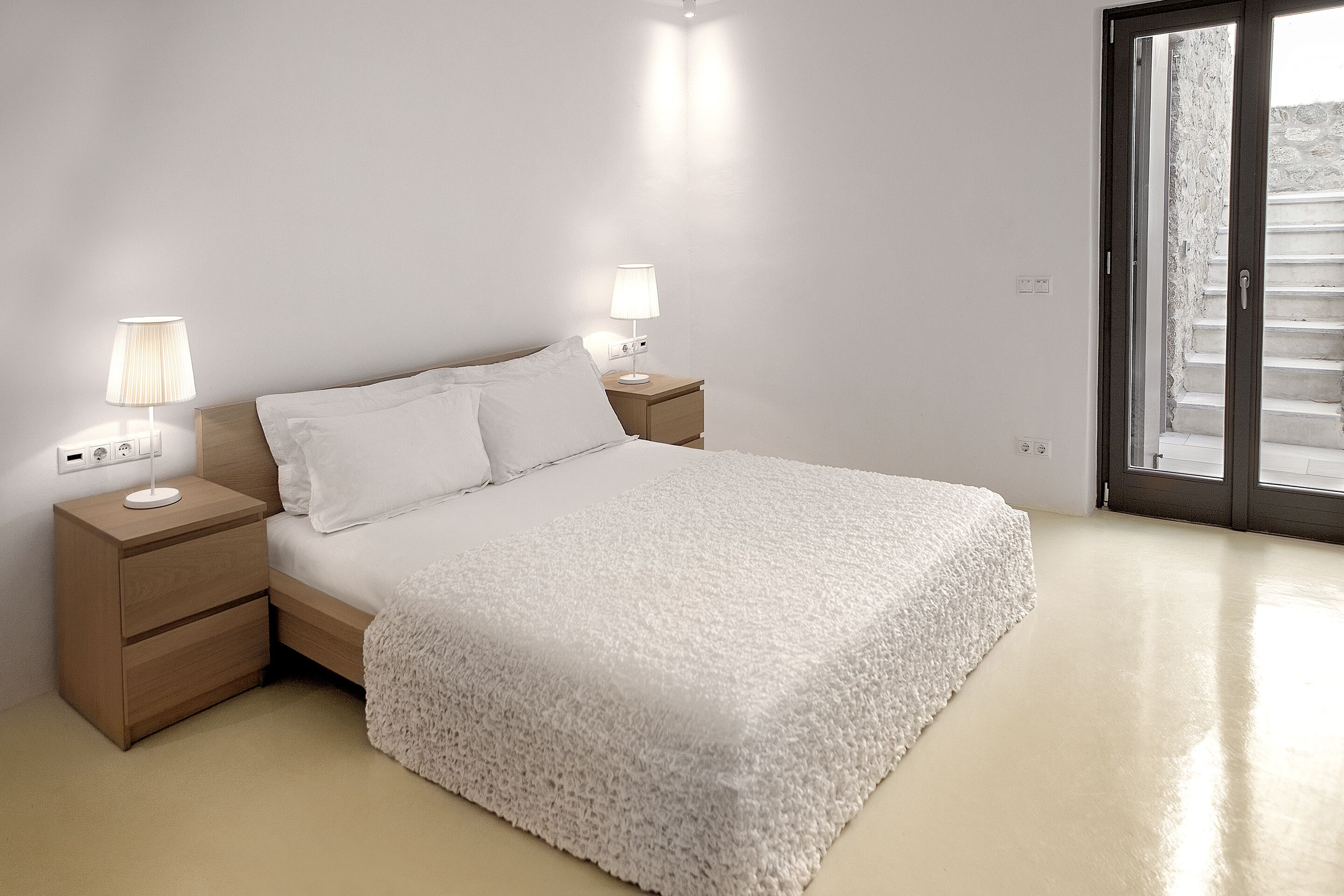 A bedroom with a large bed, two bedside tables with lamps, a wooden nightstand, and a view of a stone staircase through a glass door.
