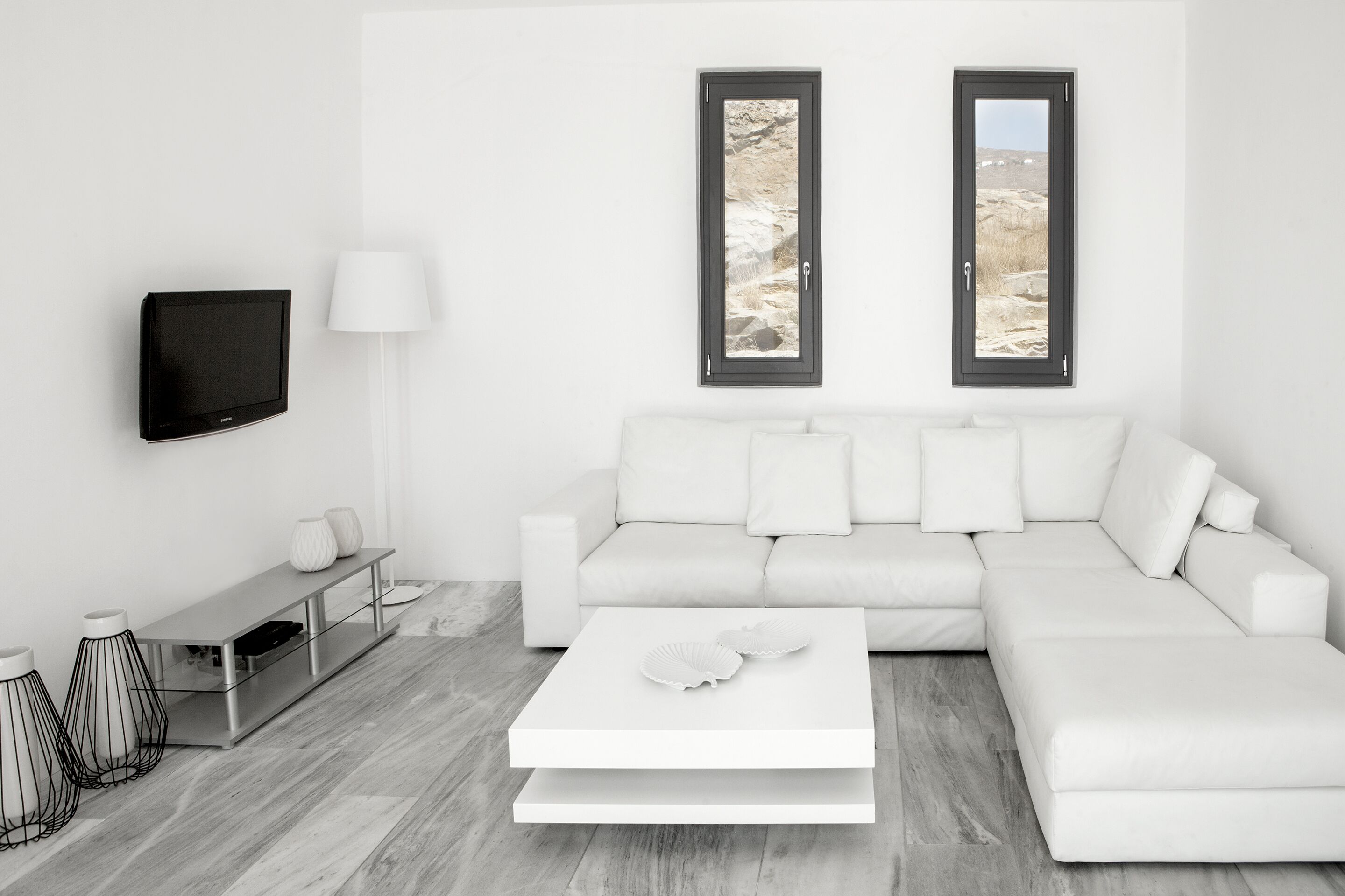 Un salon moderne avec un canapé blanc, un téléviseur à écran plat, une table basse et deux fenêtres donnant sur un paysage rocheux.