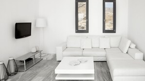 A modern living room with a white sofa, a flat-screen TV, a coffee table, and two windows with a rocky landscape view. - New Liberation Villas (Mykonos)