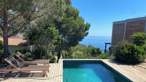 Uma piscina azul límpida com um deck de madeira e espreguiçadeiras, cercada por árvores e com vista para o mar.