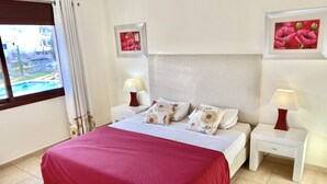 Ein Hotelzimmer mit einem Bett, zwei Nachttischen mit Lampen, einem großen Fenster mit Vorhängen und zwei gerahmten Blumenbildern an den Wänden.