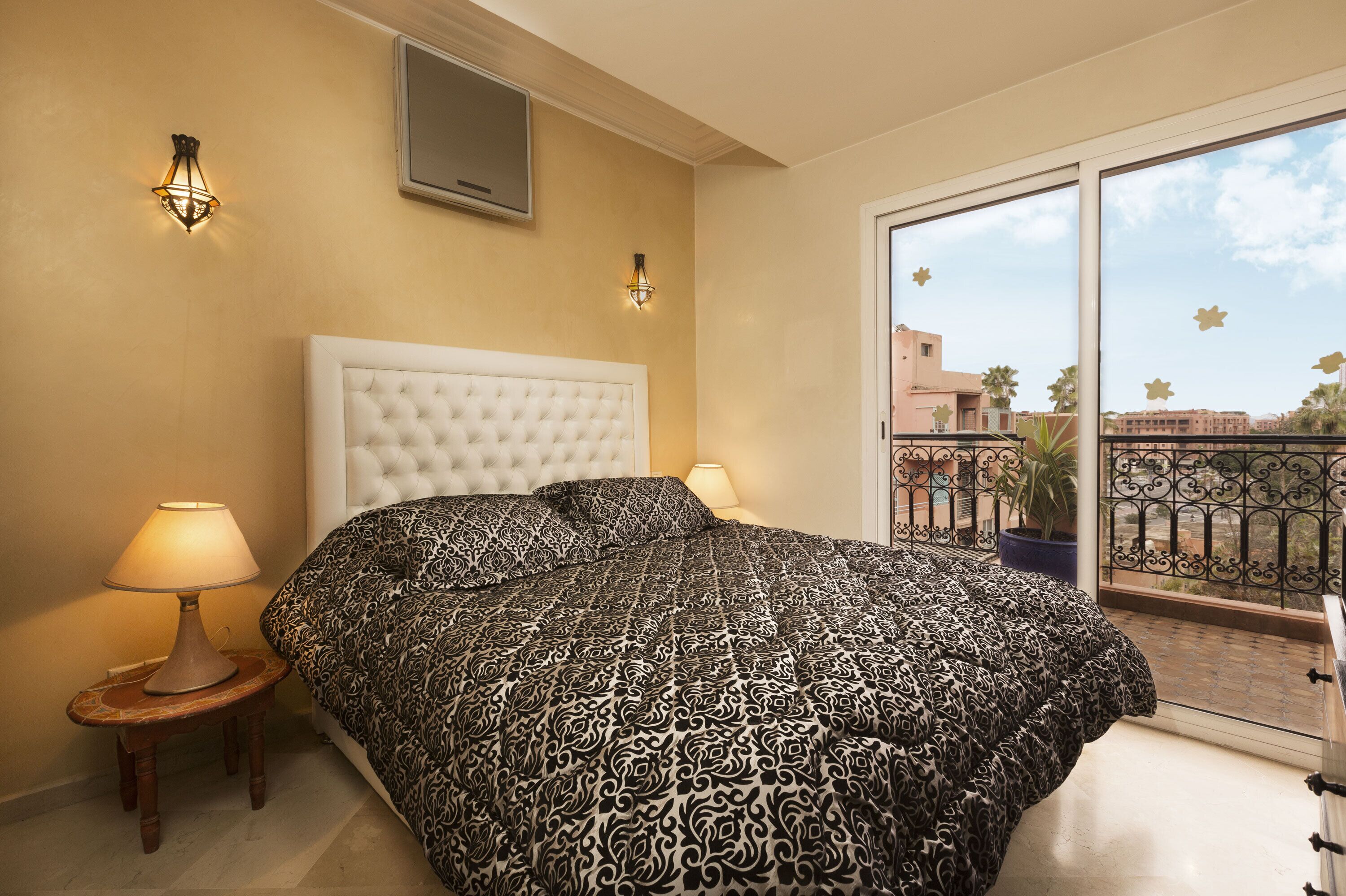 Un dormitorio con una cama grande, dos lámparas de noche, un televisor montado en la pared y un balcón con barandilla y planta en maceta.