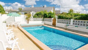 Ein rechteckiger Pool mit klarem, blauem Wasser, umgeben von einer gefliesten Liegefläche. Weiße Liegestühle und ein grüner Sonnenschirm sind vorhanden. Der Pool wird von einer Zierwand mit geometrischem Muster begrenzt.