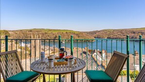 Un balcon avec une table dressée pour deux personnes, donnant sur une étendue d’eau où se trouvent des bateaux, et offrant une vue lointaine sur une ville et des collines.