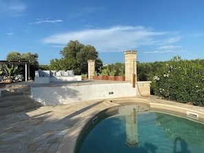 Un área de piscina con solárium de piedra, zona de estar exterior blanca y chimenea de piedra.