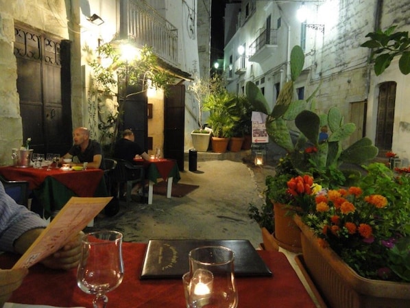 Un área de comedor al aire libre con mesas y sillas, plantas en macetas y una calle estrecha.