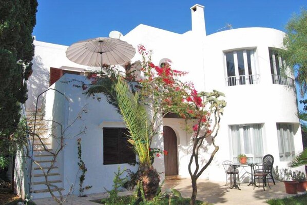Um edifício branco com área de estar coberta ao ar livre, escadaria e um pequeno jardim com plantas floridas.
